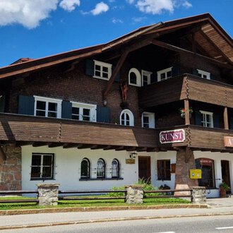 © Antik und Kunst im Malerhäusl Herbert Stumvoll Ein charmantes, traditionelles Chalet aus Holz mit mehreren Balkonen und Fenstern. Der Himmel ist blau und voller Wolken, während der grüne Rasen das Gebäude umgibt. | © Antik und Kunst im Malerhäusl Herbert Stumvoll