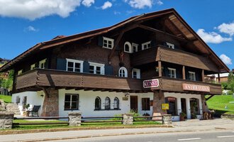 © Antik und Kunst im Malerhäusl Herbert Stumvoll Ein charmantes, traditionelles Chalet aus Holz mit mehreren Balkonen und Fenstern. Der Himmel ist blau und voller Wolken, während der grüne Rasen das Gebäude umgibt. | © Antik und Kunst im Malerhäusl Herbert Stumvoll
