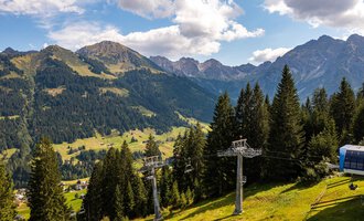 A picturesque mountain landscape with green meadows and tall fir trees. In the background, majestic mountains and a ski lift can be seen. | © OBERSTDORF · KLEINWALSERTAL BERGBAHNEN