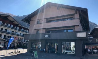 © Sport Hilbrand OG Ein modernes Gebäude mit einem Geschäft im Erdgeschoss. Die Sonne scheint auf die Fassade, und im Hintergrund sind weitere Häuser und Berge zu sehen. | © Sport Hilbrand OG