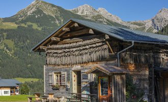 Ein traditionelles Holzhaus in den Alpen mit einem grünen Landschaftshintergrund. Die Berge sind klar und sonnig sichtbar. | © Hoflaada | Dagmar Hilbrand