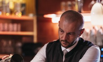 © Naturhotel Chesa Valisa | Heldentheater Ein Barkeeper schenkt einen Drink in ein Glas ein. Im Hintergrund sind Gläser und eine stilvolle Bar zu sehen. | © Naturhotel Chesa Valisa | Heldentheater