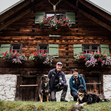 Ein rustikales Holzhaus mit blühenden Blumenkästen. Ein Paar mit zwei Hunden posiert davor. | © Hundewelt Kleinwalsertal | Stefan Lindbauer