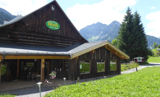 A rustic wooden building with a green sign. In the background, there are tall mountains and a blue sky. | © iKuh Schöne Ideen | Daniela Neumann