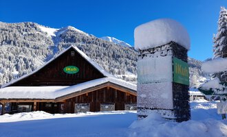 A cozy wooden house in the snow with a clear blue sky in the background. The surroundings are surrounded by snow-covered mountains and forests.
