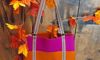 A stylish bag in orange and pink hangs in front of a rustic wooden surface. Red and orange leaves frame the image, creating an autumnal atmosphere. | © iKuh Schöne Ideen | Daniela Neumann