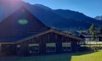 A wooden building with a sign "iKuh" in a green meadow. In the background, mountains and a clear blue sky are visible. | © iKuh Schöne Ideen | Daniela Neumann