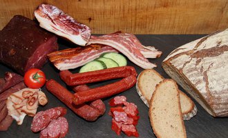 A selection of sausage and meat on a black background, accompanied by slices of bread and fresh vegetables. The arrangement is appetizing and rustic. | © Innere Wiesalpe | Andrea Kainz zuckerschnecke.at