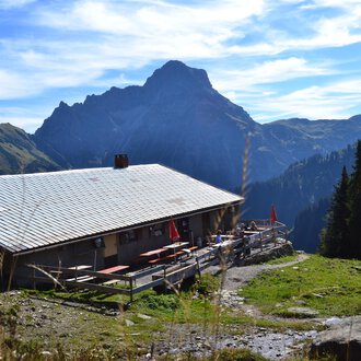 Eine alpine Hütte umgeben von Bergen und Wäldern. Der Himmel ist klar mit ein paar Wolken. | © Kleinwalsertal Tourismus