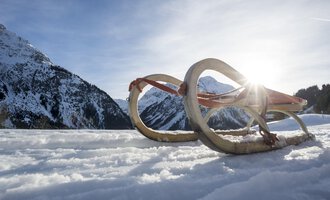 Ein Schlitten liegt im Schnee mit Bergen im Hintergrund. Die Sonne scheint sanft hinter dem Schlitten. | © Kleinwalsertal Tourismus | Dominik Berchtold