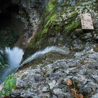 Eine felsige Klippe mit fließendem Wasser, das in einen tiefen, dunklen Pool mündet. Grüne Moose und Pflanzen wachsen zwischen den Steinen. | © Kleinwalsertal Tourismus | Andre Tappe