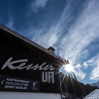 Ein Gebäude mit dem Schriftzug "Kessler" und einer Sonnenstrahlung im Hintergrund. Der Himmel ist blau mit einigen Wolken und die Umgebung wirkt alpin. | © Sport Kessler GmbH