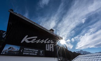 Ein Gebäude mit dem Schriftzug "Kessler" und einer Sonnenstrahlung im Hintergrund. Der Himmel ist blau mit einigen Wolken und die Umgebung wirkt alpin. | © Sport Kessler GmbH