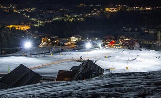 Eine verschneite Skipiste bei Nacht mit beleuchteten Liften. Im Hintergrund sind die Lichter eines kleinen Dorfes sichtbar. | © Sport Kessler GmbH