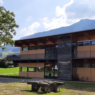© Kleinwalsertal Tourismus | Veronika Senn Ein modernes Gebäude mit großen Fenstern und Holzverkleidung. Im Vordergrund steht ein Steintisch auf einer grünen Wiese mit Bergen im Hintergrund. | © Kleinwalsertal Tourismus | Veronika Senn