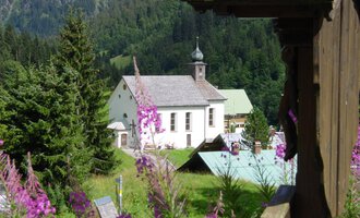 Eine malerische Landschaft mit einer Kirche und bunten Blumen im Vordergrund. Hohe Bäume und sanfte Hügel umgeben die Szene. | © Kleinwalsertal Tourismus | Rolf Köberle