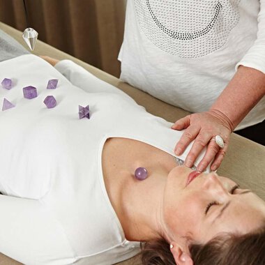 A woman is lying relaxed on a chaise lounge, while another person is placing healing stones on her body. The environment is calm and cozy. | © Bio-Hotel Oswalda Hus | Joachim Weiler