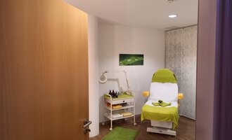 © Schönzeit | Sebastian Riezler A bright, inviting room with a treatment chair and a small table for cosmetic products. The floor is made of wood and a green carpet creates a relaxed atmosphere. | © Schönzeit | Sebastian Riezler