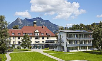 Ein charmantes Hotelgebäude mit rotem Dach und moderner Architektur. Im Hintergrund sind Berge und ein blauer Himmel zu sehen. | © Klinikverbund Allgäu