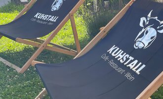 Two lounge chairs with the "Kuhstall" logo are standing on a meadow. In the background, some buildings and a welcome sign are visible. | © Kuhstall | Uwe Nagel
