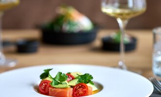 A stylish dish on a white plate, decorated with fresh tomatoes and herbs. In the background, wine glasses and another dish can be seen. | © La Vallée | Annette Sandner