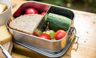 A lunchbox with fresh bread, cucumber, cherry tomatoes, grapes, and radishes. It is on a wooden surface in a natural setting. | © Kleinwalsertal Tourismus | Bastian Morell