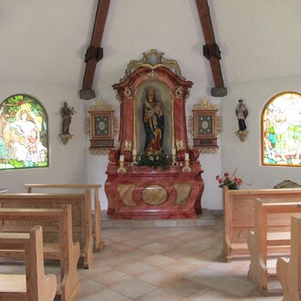Eine kleine Kapelle mit Holzbankreihen und bunten Fenstern. Im Vordergrund steht ein schmuckvoller Altar mit einer Statue. | © Stefan Heim