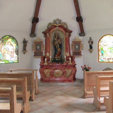Eine kleine Kapelle mit Holzbankreihen und bunten Fenstern. Im Vordergrund steht ein schmuckvoller Altar mit einer Statue. | © Stefan Heim
