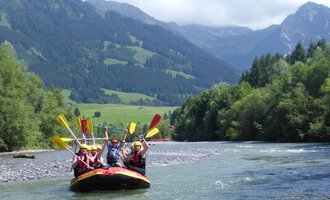 A group of people is paddling in an inflatable boat on a river. The surrounding landscape is green and mountainous, with a clear sky in the background. | © MAP-Erlebnis