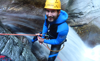 A man in a blue neoprene suit is climbing at a waterfall. He is wearing a helmet and is secured with a rope. | © MAP-Erlebnis