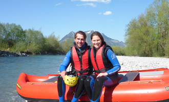 A couple is sitting and smiling by a riverbank in water rafting gear. In the background, trees and mountains can be seen. | © MAP-Erlebnis