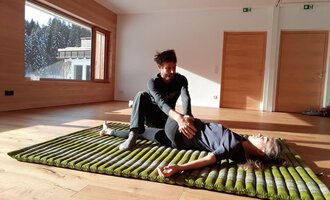 Eine Person liegt auf einer Matte, während eine andere Person sie sanft behandelt. Der Raum hat große Fenster mit Blick auf Bäume und viel Tageslicht. | © Alexander Mayr