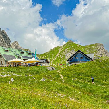 A picturesque mountain landscape with cozy cottages and green meadows. In the background, impressive rocks and a blue sky with clouds can be seen. | © Mindelheimer Hütte | Wolfgang Stache
