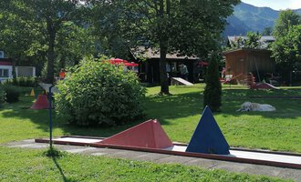 Ein Minigolfplatz mit bunten Hindernissen in einer grünen Umgebung. Im Hintergrund sind Bäume und ein Gebäudeteil zu sehen. | © Kleinwalsertal Tourismus | N. Lughammer