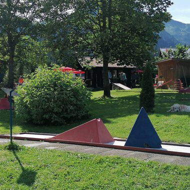 Ein Minigolfplatz mit bunten Hindernissen in einer grünen Umgebung. Im Hintergrund sind Bäume und ein Gebäudeteil zu sehen. | © Kleinwalsertal Tourismus | N. Lughammer
