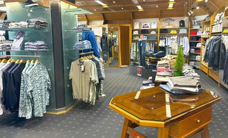 © Moosbrugger Mode e.U. Ein moderner Bekleidungsladen mit verschiedenen Shirts und Hosen. Im Vordergrund steht ein Holztisch, während die Regale im Hintergrund voll mit Kleidung sind. | © Moosbrugger Mode e.U.