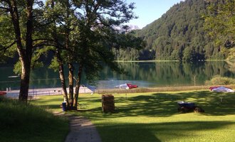 © Kleinwalsertal Tourismus | Naturbad Freibergsee Ein ruhiger See umgeben von Bäumen und grünen Wiesen. Im Hintergrund sind Berge und Sonnenliegen am Ufer zu sehen. | © Kleinwalsertal Tourismus | Naturbad Freibergsee