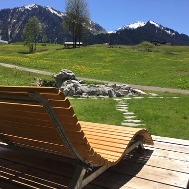 Eine Liege aus Holz steht auf einer Terrasse mit Blick auf grüne Wiesen und schneebedeckte Berge im Hintergrund. Der Himmel ist klar und die Landschaft wirkt idyllisch. | © Kleinwalsertal Tourismus | Sarina Berchtold