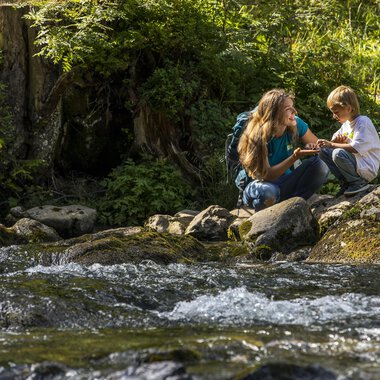 A mother and her son are sitting by a clear stream in nature. They are having fun and enjoying their time together. | © Kleinwalsertal Tourismus | Frank Drechsel