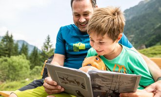 A father and his son are sitting outside and reading a book together. Both are smiling and the boy is holding a stuffed animal in his hand. | © Kleinwalsertal Tourismus | Bastian Morell