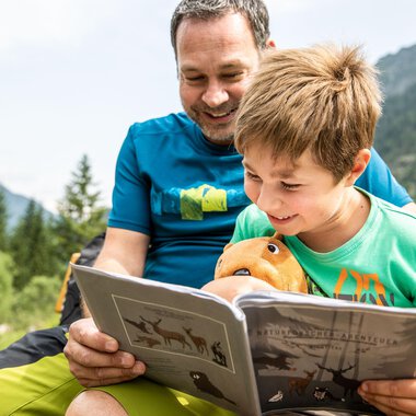 Ein Vater und sein Sohn sitzen im Freien und lesen gemeinsam ein Buch. Beide lächeln und der Junge hält ein Stofftier in der Hand. | © Kleinwalsertal Tourismus | Bastian Morell