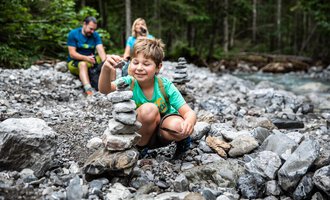 A boy is building a small stone tower by the riverbank. In the background, there are two adults enjoying nature. | © Kleinwalsertal Tourismus | Bastian Morell