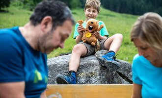 A boy sits on a rock holding a plush toy lion in his hand. In the background, two adults are busy, while a green meadow and trees can be seen. | © Kleinwalsertal Tourismus | Bastian Morell