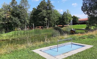A small swimming pool in a green meadow, surrounded by trees. In the background, playground equipment and a building can be seen. | © Kleinwalsertal Tourismus | Veronika Senn