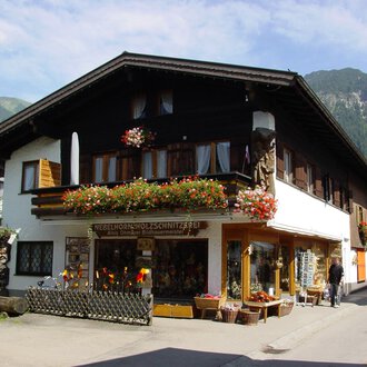 © Nebelhorn Holzschnitzerei e.K. Ein traditionelles Haus in den Bergen, umgeben von Grün. Der Laden im Erdgeschoss bietet handgefertigte Holzschnitzereien an und ist mit Blumen geschmückt. | © Nebelhorn Holzschnitzerei e.K.