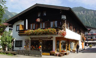 © Nebelhorn Holzschnitzerei e.K. Ein traditionelles Haus in den Bergen, umgeben von Grün. Der Laden im Erdgeschoss bietet handgefertigte Holzschnitzereien an und ist mit Blumen geschmückt. | © Nebelhorn Holzschnitzerei e.K.