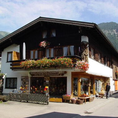 Ein traditionelles Haus in den Bergen, umgeben von Grün. Der Laden im Erdgeschoss bietet handgefertigte Holzschnitzereien an und ist mit Blumen geschmückt. | © Nebelhorn Holzschnitzerei e.K.
