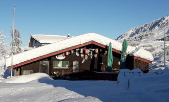A cozy cabin in the snow, surrounded by a wintry landscape. The sky is clear and the mountains are clearly visible.