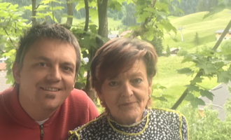 A man and a woman are standing together in nature. In the background, green hills and trees can be seen.