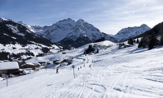 © OBERSTDORF · KLEINWALSERTAL BERGBAHNEN Eine verschneite Landschaft mit Bergen im Hintergrund. Skifahrer genießen die Pisten in dieser ruhigen Winterszene. | © OBERSTDORF · KLEINWALSERTAL BERGBAHNEN